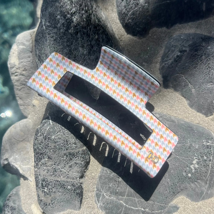 A rectangular hair claw clip with a pastel orange, yellow, and white gingham pattern, resting on a textured background of dark volcanic rocks and sand.