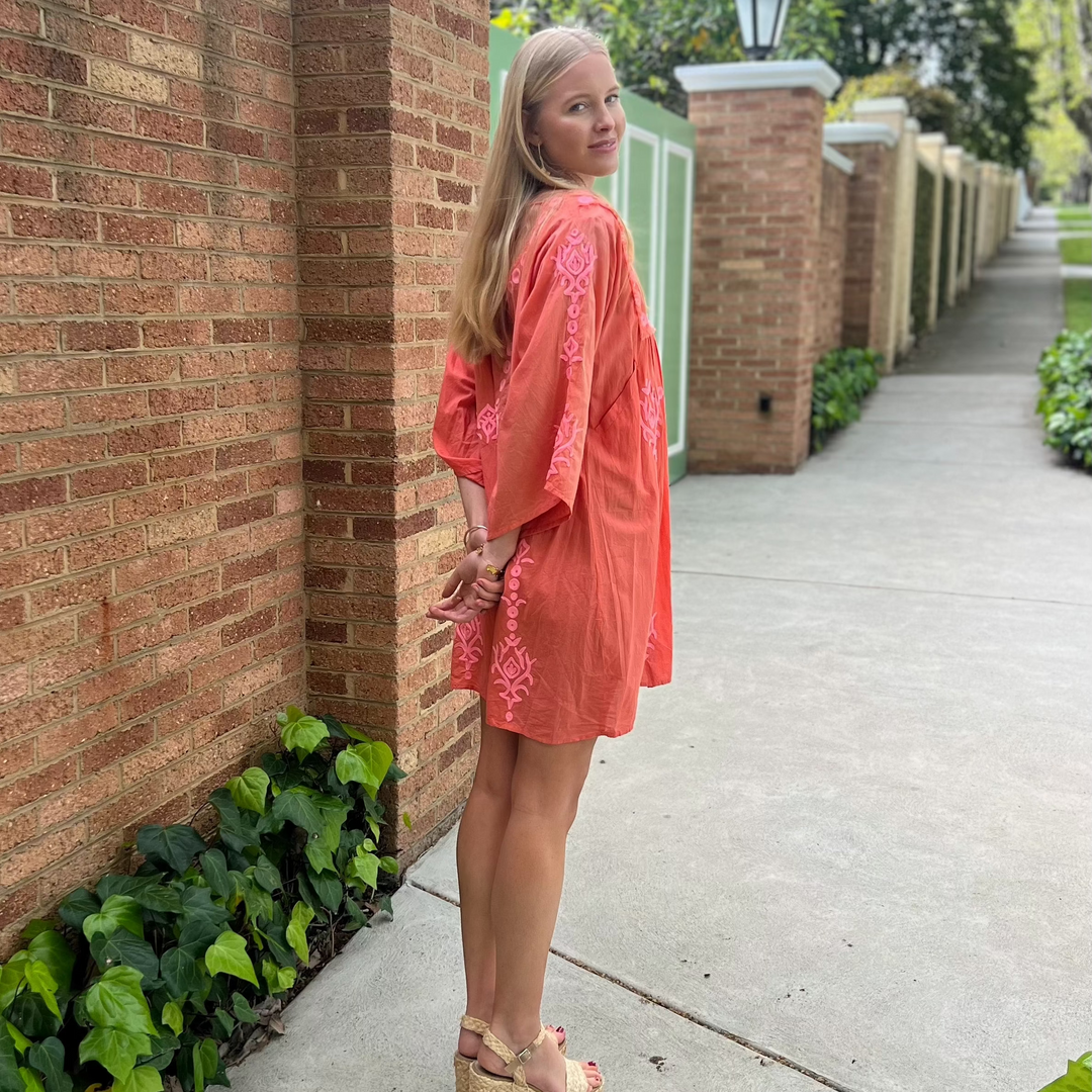 Woman in a coral dress standing against a brick wall on a sidewalk.
