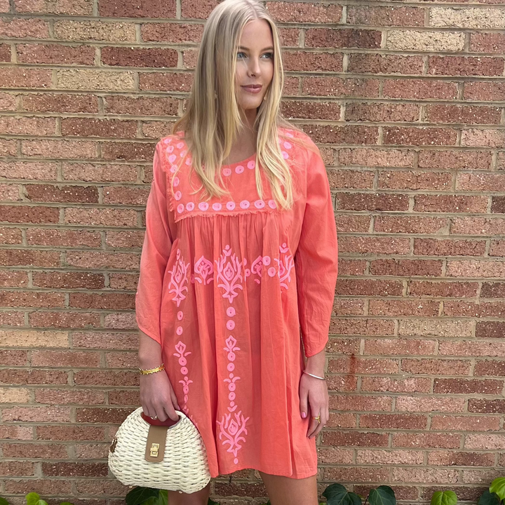Woman wearing a coral dress with pink embroidery against a brick wall.