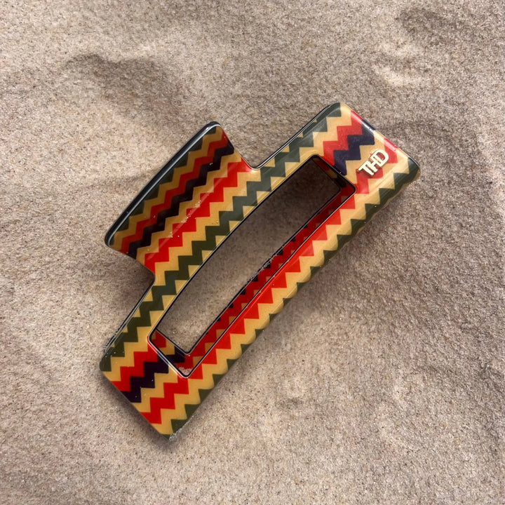 A rectangular hair claw clip with a colorful chevron pattern of red, yellow, green, and black, resting on a textured beige fabric background.